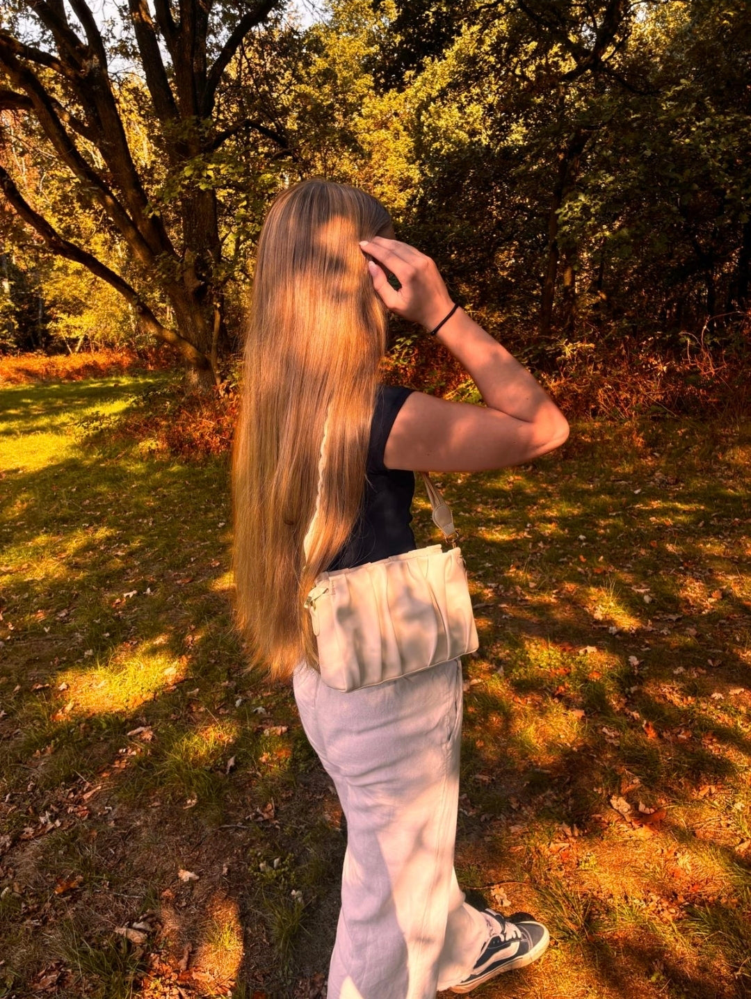 Frau mit langen blonden Haaren trägt die Avyline Aelyvre Pearl Handtasche in hellem Cremeweiß bei einem Spaziergang durch einen herbstlichen Wald.